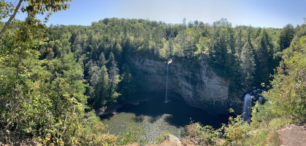 A tall waterfall dropping into a forested canyon pool, seen from a trail overlook by Adam Neuerburg