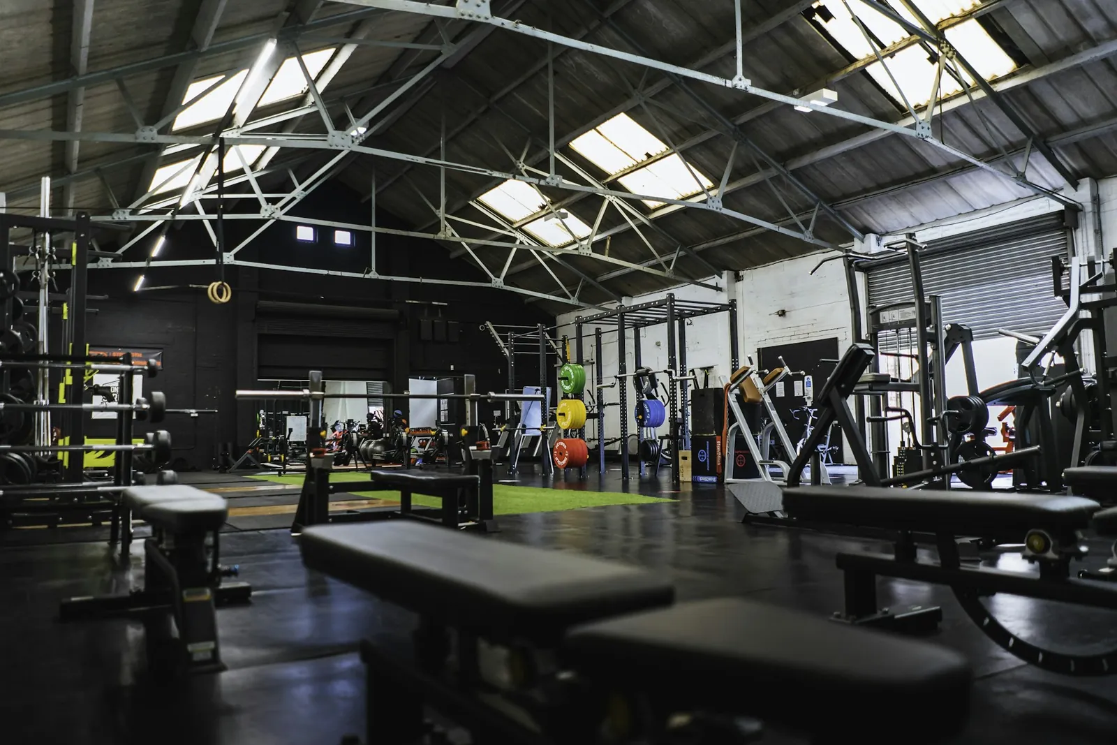 Wide view of a warehouse-style gym floor with benches, racks and skylights