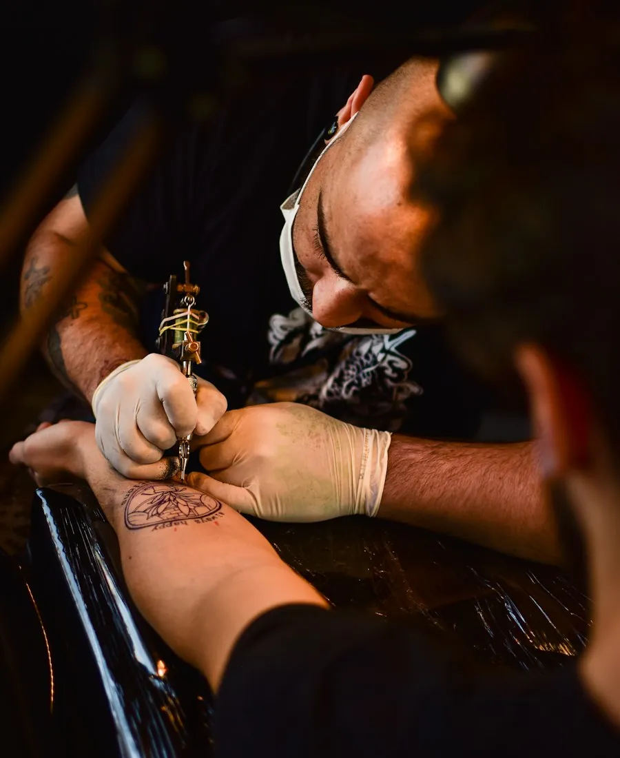 Tattoo artist leaning in over a client&rsquo;s arm under a work lamp