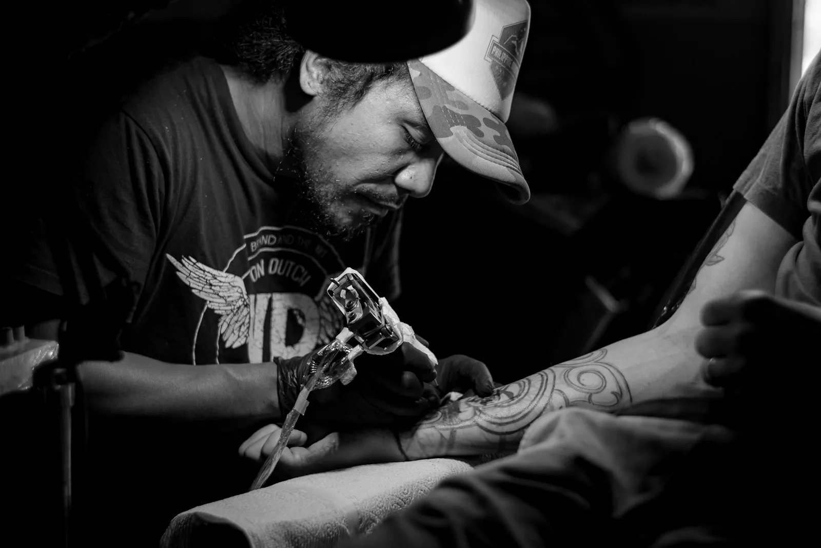 Tattoo artist in a cap bent over a client&rsquo;s forearm, needle in hand